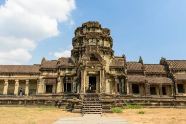 Angkor Wat 'ın yıpranmış kumtaşı giriş köşkü açık güneş ışığında duruyor, kuru çimenler ve açık mavi bir gökyüzü tarafından çerçevelenmiş. Karmaşık oymalar ve simetrik çizgiler bu ikonik Kamboçya tapınağının muazzam Khmer mimarisini vurguluyor..