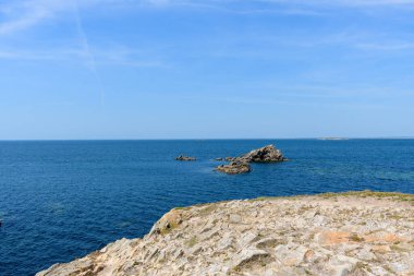Sivri kayalar, Saint Pierre Quiberon 'daki Pointe Beg-An-Aud' da güneşli bir kıyı şeridinden derin mavi okyanusa doğru uzanır, berrak gökyüzü ve sakin suyla huzurlu bir deniz manzarası yaratır..