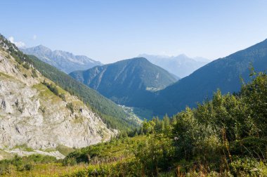Sarp kayalıklar ve ormanlık tepelerle çevrili yeşil bir alp vadisi üzerinde geniş bir manzara berrak mavi gökyüzü ve manzarayı aydınlatan yumuşak yaz ışığı.