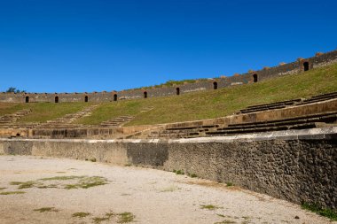 Pompeii, İtalya 'daki Roma amfitiyatrosu içindeki eğimli katlarda antik taş koltuklar yükselir. Parlak güneş ışığı, tarihi eserleri vurgulayarak, canlı mavi gökyüzünün altında dokulu volkanik duvarları ve çimenli setleri vurguluyor.
