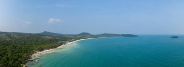 Geniş panoramik manzara, Kamboçya 'nın Koh Rong adasındaki Long Set Sahili' nin beyaz kumla turkuaz denizin birleştiği geniş eğrisini gösteriyor. Bereketli orman ve yumuşak tepeler berrak mavi gökyüzünün altındaki sakin kıyı şeridini çevrelemektedir..
