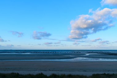 Utah Sahili 'nin geniş manzarası sakin gel-git havuzları, yumuşak kum ve açık mavi gökyüzünde süzülen yumuşak bulutlar. İnce ışık, sakin kıyı şeridini ve açık kıyı atmosferini vurguluyor.