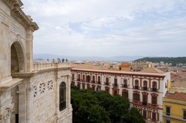 Bastione di Saint Remy 'nin detaylı taş işi yapraklı ağaçların ve tarihi binaların üzerinde yükselir, Cagliaris şehir manzarası ve bulutlu gökyüzünün altındaki uzak tepeler.