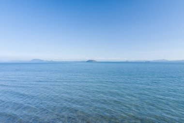 Geniş, berrak mavi sular Nagasaki yakınlarında parlak bir gökyüzü altında uzak bir ada ve dağlara doğru uzanır. Sakin deniz manzarası yumuşak güneş ışığıyla yıkanır, dingin ve açık bir atmosfer yaratır..