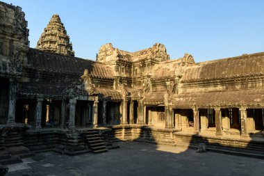 Sıcak öğleden sonra güneş ışığı, yıpranmış kumtaşı galerileri boyunca gölgeler bıraktı ve Kamboçya 'nın Angkor Vat zirvesinde avluya çıktı. Karmaşık çatı çizgileri ve antik Khmer mimari detayları berrak mavi bir gökyüzünün altında göze çarpıyor..