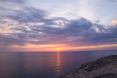 Parlak turuncu güneş sakin bir denizin üzerinde batar, sıcak yansımalar oluştururken dramatik bulutlar engebeli kayalıkların üzerinde sürüklenir. Huzurlu manzara, Stintino yakınlarındaki Sardunya kıyılarının doğal güzelliklerini yansıtıyor..