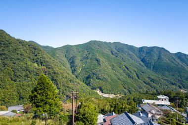 Yoğun ormanlık dağların eteklerindeki küçük bir köy yuvasının geleneksel çatıları berrak mavi bir gökyüzünün altında. Güneş ışığı canlı yeşilliği ve yuvarlanan manzarayı vurguluyor, huzurlu bir kırsal manzara yaratıyor..