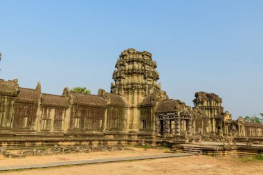 Kamboçya 'daki Angkor Vat tapınağının üzerinde berrak mavi bir gökyüzünün altında yıpranmış bir kumtaşı kulesi yükseliyor. Antik Khmer mimarisi karmaşık oymalarla ve kuru otlarla çevrili, tarih ve tropikal bir duyguyu çağrıştırıyor.