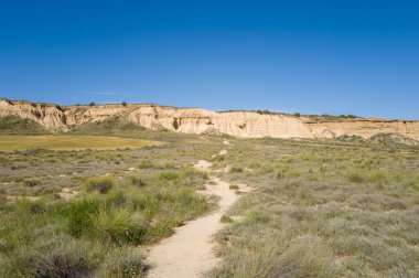 Sarmal bir toprak yol, İspanya 'nın Bardenas Reales kentindeki parlak mavi gökyüzünün altındaki engebeli bej kayalıklara doğru yeşil çalıları ve kuru otları kesiyor. Açık arazi vahşi, güneşli ve geniş hissettiriyor..