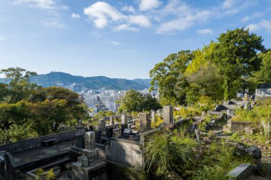 Mezar taşları ve yemyeşil yeşillikler, Japonya 'nın Nagasaki kentini, uzak dağları ve parlak mavi gökyüzünün altındaki kentsel ufuk çizgisini gösteren panoramik bir manzara sunar. Güneş ışığı, taşların ve yaprakların dokusunu vurgular, barışçıl ve yükseltilmiş bir yapı yaratır.