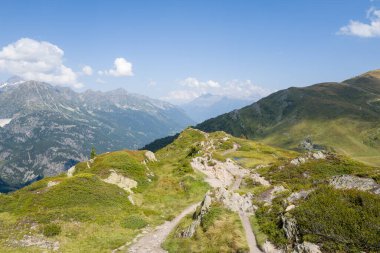 Kaya ve alçak çalılarla benekli çimenli bir tepe boyunca daralan yürüyüş yolu Aiguillettes des Posettes bölgesinde açık mavi bir gökyüzü altında engebeli tepeler ve derin vadiler sunuyor..