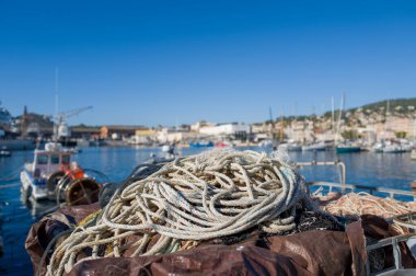 La Ciotat limanında demirli bir teknede bulunan sarmalanmış halat ve ağların yakın çekimi, güneşli bir marina, mavi gökyüzü ve arka planda bulanık deniz manzaralı binalar. Sahne, deniz dokularını ve rahat bir sahil atmosferini ön plana çıkarıyor..