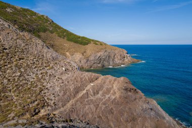 İki keçi, Arjantin yakınlarındaki canlı mavi Akdeniz 'in üzerindeki sarp, dokulu uçurumlarda duruyor. Vahşi kıyı şeridinde kayalık oluşumlar, seyrek bitki örtüsü ve berrak bir gökyüzünün altında berrak su bulunur..