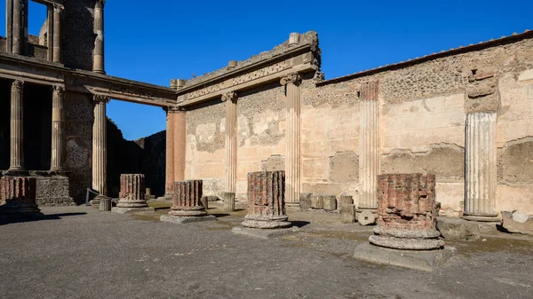 Taş sütunlar ve yıpranmış duvarcılık, İtalya 'nın Pompeii arkeoloji sahasındaki bazilikanın büyük cephesini oluşturur. Parlak güneş ışığı, canlı mavi gökyüzünün altında klasik Roma mimarisini ve dokulu yüzeyleri ortaya çıkarıyor..