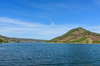 Yeşil ve kırmızı tepe, Salagou Gölü 'nün parıldayan yüzeyinin üzerinde dokulu suyla ve açık gökyüzüyle barışçıl bir doğal manzara yaratarak yükselir..