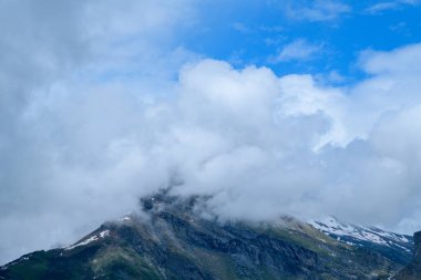 Alçak asılı bulutlar dağlık bir dağın tepesinde sürükleniyor. Dağılmış kar parçaları ve yukarıdaki canlı mavi gökyüzü Fransız Alplerinde dramatik ve kasvetli bir alp manzarası yaratıyor..