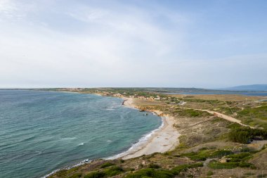 Sardinya, Capo San Marco yakınlarındaki turkuvaz suları boyunca kıvrılan kumlu bir kıyı şeridinin havadan görünüşü, kumullar ve Akdeniz bitkileri arasında uzanan küçük bir kıyı köyü, parlak, berrak bir gökyüzü altında..
