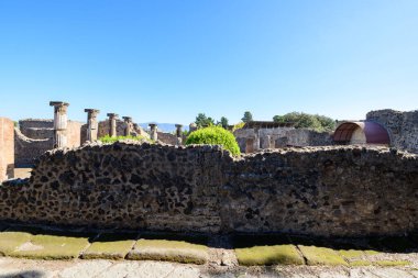 Yıpranmış volkanik taş duvarlar ve antik Roma sütunları İtalya 'nın Pompeii kentindeki arkeolojik bölgede canlı bir mavi gökyüzünün altında duruyor. Güneş ışığı, tarihi kalıntıların ötesindeki pürüzlü duvar dokularını, yeşil çalıları ve uzak dağları vurguluyor..