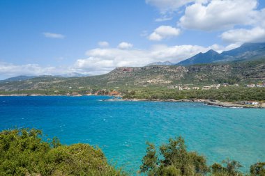 Berrak turkuaz su kıvrımları dağınık evleri ve yemyeşil tepeleri olan engebeli bir kıyı şeridi boyunca beyaz bulutlarla kaplı parlak bir gökyüzünün altında. Dağlık zemin ve doğal bitki örtüsü canlı bir Akdeniz yaz manzarası yaratır..