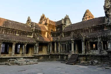 Ilık güneş ışığı, Angkor Vat, Kamboçya 'daki yıpranmış kumtaşı duvarları ve iç galerinin sütunlarını otlatıyor. Tarihi Khmer tapınağı mimarisi dokulu taş yüzeyler ve açık mavi gökyüzü ile çerçevelenmiştir..
