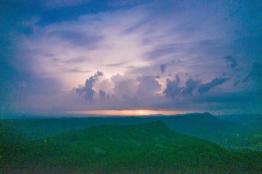 Ufukta yükselen gök gürültülü bulutların altında tek bir yıldırım çakar ve alacakaranlıkta karanlık bir dağ manzarasını aydınlatır. Sahnede katmanlı siluetler, parlayan gökyüzü ve kasvetli bir alacakaranlık atmosferi yer alıyor..