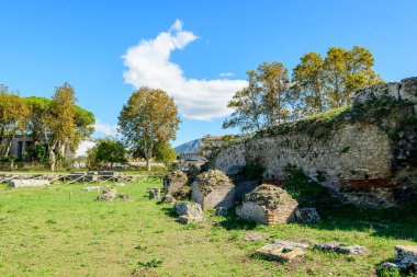 Antik taş ve tuğla ustalığı, İtalya 'nın Paestum kentindeki çimenli arkeolojik alanlarla çevrili yıpranmış bir duvar oluşturur. Arazi olgun ağaçlar tarafından çerçevelenmiş ve parlak bir mavi Akdeniz gökyüzünün altında parlak güneş ışığı ile aydınlatılmıştır..