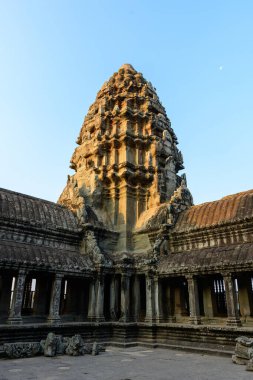 Angkor Vat 'ın ana kumtaşı kulesi yumuşak akşam güneşinde parlar, yıpranmış galerilerin ve karmaşık sütunların üzerinde yükselir. Açık mavi gökyüzü ve ince gölgeler Khmer mimarisini ve tarihi taş işçiliğini vurguluyor..