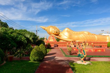 Manikürlü bahçeler ve Vientiane, Laos 'taki Pha That Luang' daki tropikal yeşilliklerle çevrili süslü kırmızı bir heykel üzerinde uzanan büyük altın bir Buda heykeli. Parlak güneş ışığı, pürüzsüz hatları ve karşı dekoratif ayrıntıları vurgular