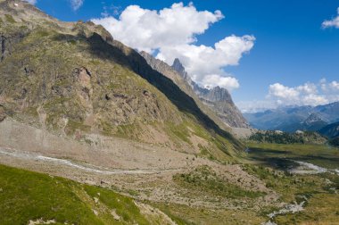 Col de la Seigne yakınlarındaki İtalyan Alp Dağları 'ndaki yeşil bir vadide yükselen kayalık kayalıklar, dolambaçlı bir dere ve dağınık bulutlarla dolu parlak mavi bir gökyüzünün altında dramatik tepeler..