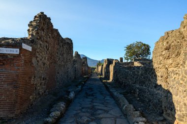 Pompeii, İtalya 'nın yıkıntıları arasında volkanik taş ve tuğla duvarlarla çevrili uzun düz Roma caddesi. Erken güneş ışığı derin gölgeler oluştururken, Vezüv Dağı açık mavi gökyüzünün altında yükselir..