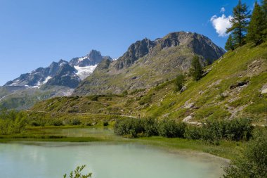 Soluk turkuvaz nehri, dramatik kayalık dağların eteklerindeki yemyeşil bitki örtüsü boyunca uzanır. Kar yığınları ve açık mavi gökyüzü..