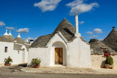 Konik taştan çatısı ve kemerli girişi olan tek bir geleneksel trullo evi Alberobello, Apulia, İtalya 'da parlak mavi bir gökyüzünün altında duruyor. Beyazlatılmış duvarlar, kırsal ahşap kapılar ve saksı bitkileri Akdeniz mimari tarzını ön plana çıkarıyor