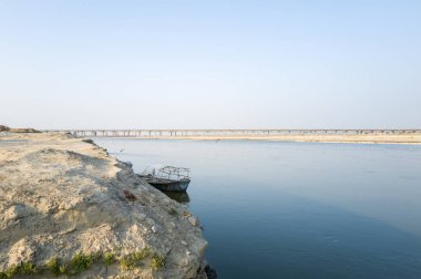 Hindistan 'ın Prayagraj kenti yakınlarında kumlu seti ve teknesi olan geniş bir nehrin minimalist manzarası. Uzaktaki uzun köprü berrak mavi bir gökyüzünün altındaki suyu gerer..