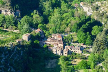 Navacelles 'teki yoğun yeşil orman ve kayalık yamaçların arasına yerleştirilmiş terakotta çatıları olan küçük bir köyün hava görüntüsü. Parlak gün ışığı, geleneksel binaların canlı yapraklarını ve kırsal dokularını güçlendirir..