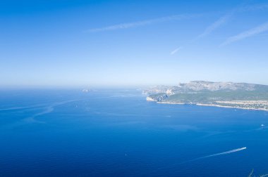 Sakin mavi Akdeniz suları üzerinde geniş bir manzara engebeli Calanques uçurumları ve bulutsuz bir gökyüzünün altında kıyı şeridi boyunca uzanan yeşil tepeler. Sahne açık, huzurlu ve güneşli, suda ince tekne izleri var..