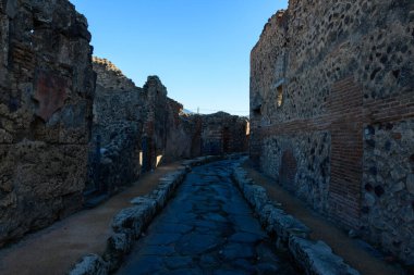 Pompeii, İtalya 'da yıpranmış volkanik taş ve tuğla duvarlarla kaplı dar bir Roma geçidi. Güneş ışığının derin gölgeleri ve lekeleri berrak mavi gökyüzünün altındaki pürüzsüz duvarları ve tarihi dokuları vurgular..