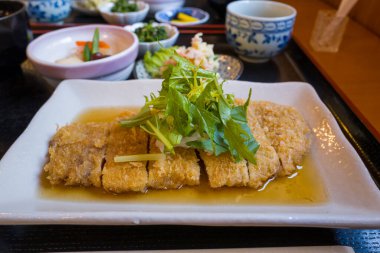 Hafif soslu, üzerinde taze yapraklı yeşillikler bulunan ve arka planda çeşitli mezeler bulunan dikdörtgen bir tabakta servis edilen domuz pirzolası. Ilık iç aydınlatma dokuları ve davet edici sunumu vurgular.