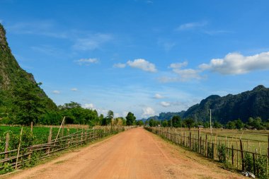 Kırsal ahşap çitlerle çevrili düz kırmızı toprak yolu yeşil tarlaları ve canlı mavi gökyüzünün altındaki uzak kireçtaşı tepeleriyle birlikte açık tarım arazisinden geçer. Manzara doğal dokulara ve Laos 'un tipik kırsal atmosferine sahiptir..