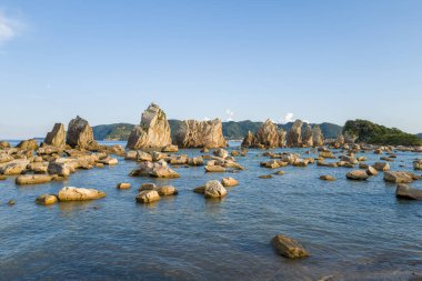 Kümelenmiş taş kuleler ve dağınık kayalar Japonya 'nın Kushimoto kentindeki Hashiguiwa' daki sığ mavi sulardan yükseliyor. Orman tepeleri ve açık bir gökyüzünün altında küçük bir ada var..