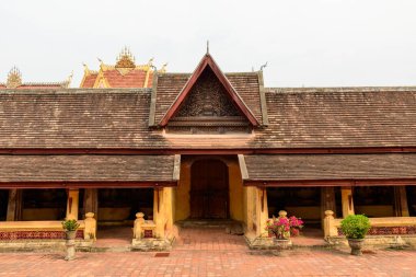 Vientiane, Laos 'taki Wat Si Saket tapınağındaki tarihi bir galerinin önünde klasik eğimli kiremitli çatı, süslü ahşap kapı ve sarı sütunlar yer alıyor. Saksı bitkileri ve tuğla döşeme yumuşak gün ışığında sakin tapınak avlusuna detay ekler.