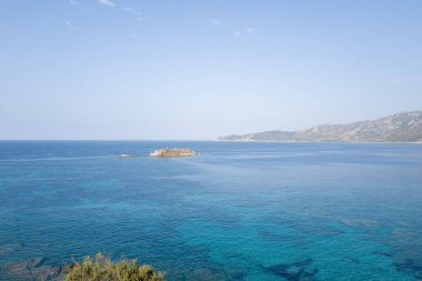 Cala Caterina açıklarında güneşli bir kayalık adanın çevresini saran berrak, berrak, mavi bir su, uzak dağları ve sakin bir Akdeniz manzarası yaratan solgun bir gökyüzünü kaplar..