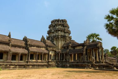 Yıpranmış kumtaşı duvarlar ve süslü oymalar Angkor Wat 'ın giriş köşkünü tanımlar, öğlen güneşiyle aydınlatılır ve kuru çimenler ve palmiye ağaçlarıyla çevrilidir. Sahne, bunun muazzam simetrisini ve karmaşık Khmer mimarisini vurguluyor.