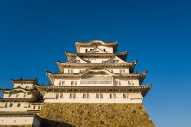 Himeji Kalesi 'nin heybetli ana kalesinin düşük açılı görüntüsü katmanlı beyaz cephelerini ve dayanıklı, mavi gökyüzüne karşı kurulmuş taş taban üzerindeki karmaşık kiremitli çatılarını gözler önüne seriyor..