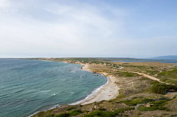 Sardinya, Capo San Marco yakınlarındaki turkuvaz suları boyunca kıvrılan kumlu bir kıyı şeridinin havadan görünüşü, kumullar ve Akdeniz bitkileri arasında uzanan küçük bir kıyı köyü, parlak, berrak bir gökyüzü altında..