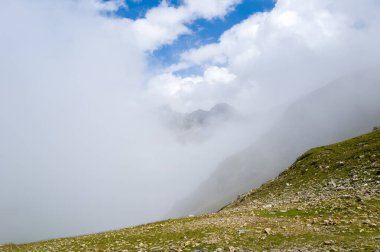 Alçak bulutlar ve sis, Col du Bonhomme 'da taşlı yeşil bir yamaçta sürükleniyor. Mavi gökyüzünün parlak bir parçasının altındaki uzak dağ zirvelerini kısmen gizliyor..