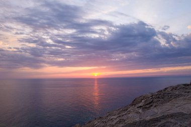 Sıcak akşam güneşi, Sardunya 'nın Stintino yakınlarındaki engebeli kayalıkların üzerinde dokulu bulutlar sürüklenirken sakin Akdeniz sularında yansıyor. Huzurlu sahne yumuşak pastel renkler ve huzurlu kıyı atmosferiyle dolu..