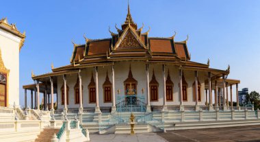 Gümüş Pagoda, Kamboçya 'nın Phnom Penh kentindeki Kraliyet Sarayı kompleksi içinde gün ışığında duruyor. Süslü altın Khmer çatı çizgileri, kırmızı ve altın pencere panelleri ve dekoratif sütunlar açık mavi gökyüzüne karşı vurgulanır, çarpıcı bir mavi gökyüzü oluşturur