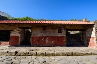 Pompeii, İtalya 'da eski bir Romalı dükkanının yıpranmış taş ve alçı cephesinde fayanslı bir çatının altında solmuş kırmızı bir duvar resmi ve Latince yazıtlar vardı. Parlak güneş ışığı, taş döşeli cadde boyunca sert dokuları ve tarihi karakteri vurguluyor.