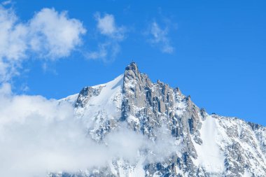Aiguille du Midi dimdik ayakta duruyor, karla kaplı sırtlarıyla sürüklenen bulutları delip geçiyor, Fransız Alpleri 'ndeki parlak mavi gökyüzüne karşı..