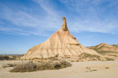 Bardenas Reales, İspanya 'nın kurak kumlu arazisinden, geniş mavi bir gökyüzünün altından çarpıcı bir bej kaya zirvesi yükselir. Güneş ışığı, engebeli ve açık bir manzarada katmanlı dokuları ve yontulmuş hatları gösterir..